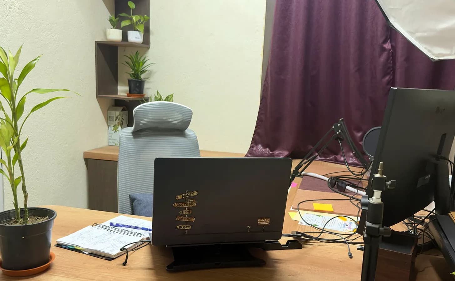 A desk with a laptop on a stand, an open notebook and a potted plant, a grey chair behind it and a softbox light by a plum curtain, Kolkata, India