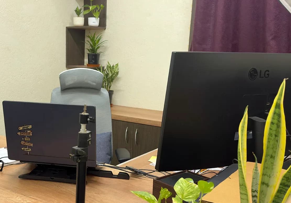 A laptop on a stand with signpost stickers on its lid, an LG monitor seen from behind, and an empty grey chair below a corner shelf of plants, Kolkata, India