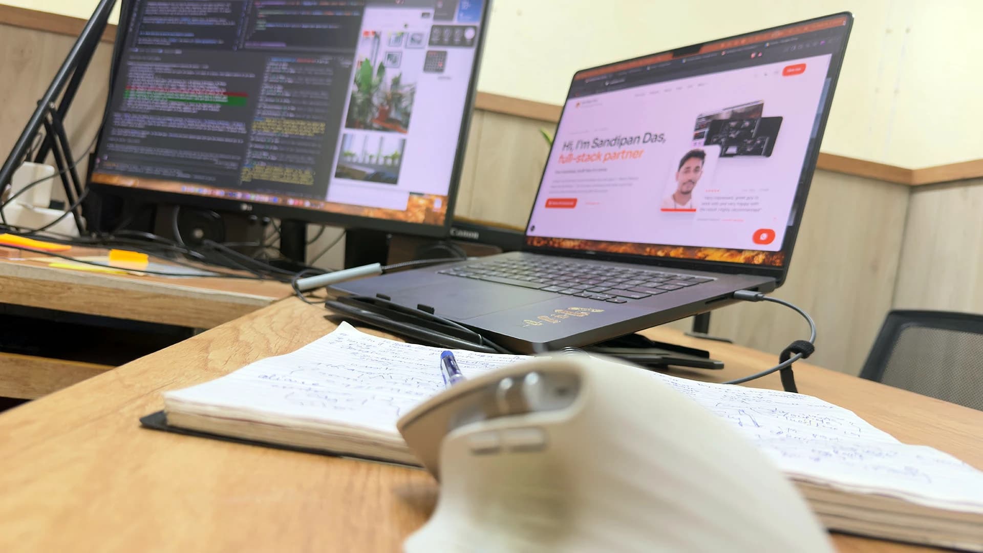 A laptop raised on a stand next to a coding monitor, with a handwritten notebook, pen and white mouse in front, Kolkata, India